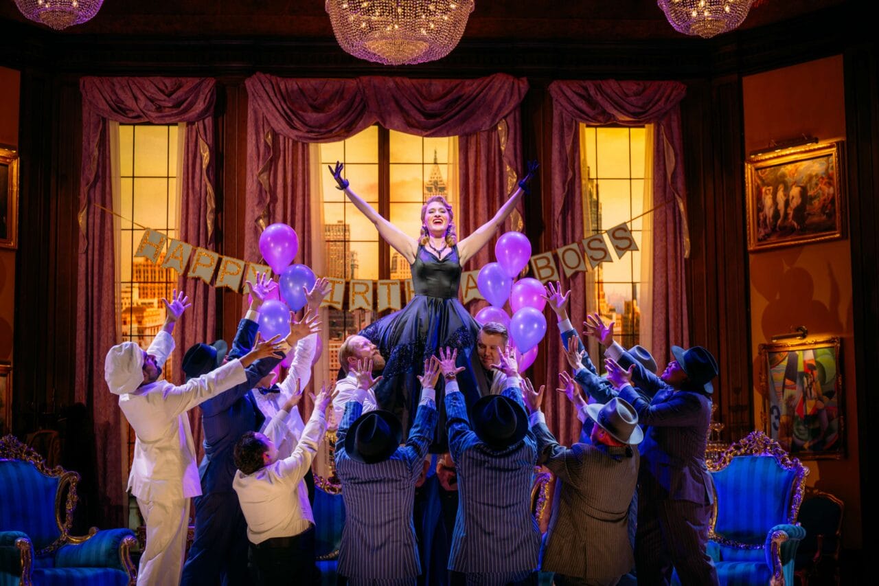 A woman in a black dress stands on a blue couch holding purple balloons, surrounded by people in suits, on a stage decorated for a party with a banner reading "HAPPY BIRTHDAY BOSS.