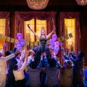 A woman in a black dress stands on a blue couch holding purple balloons, surrounded by people in suits, on a stage decorated for a party with a banner reading "HAPPY BIRTHDAY BOSS.