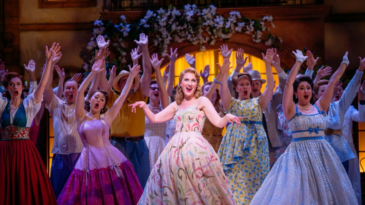 A group of performers in colorful vintage costumes sing and gesture with raised arms on a brightly lit stage decorated with flowers.