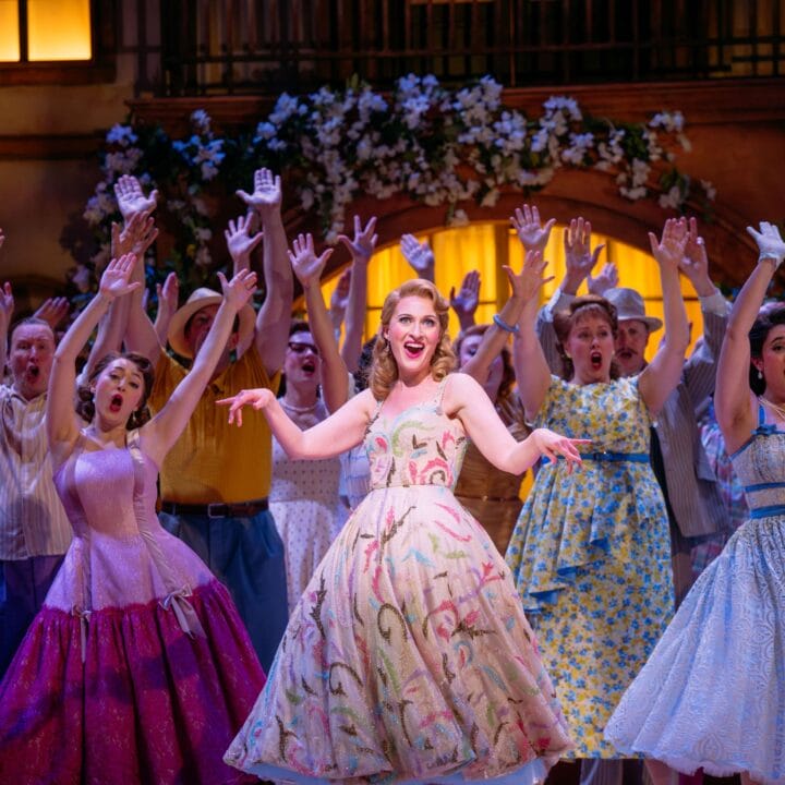 A group of performers in colorful vintage costumes sing and gesture with raised arms on a brightly lit stage decorated with flowers.