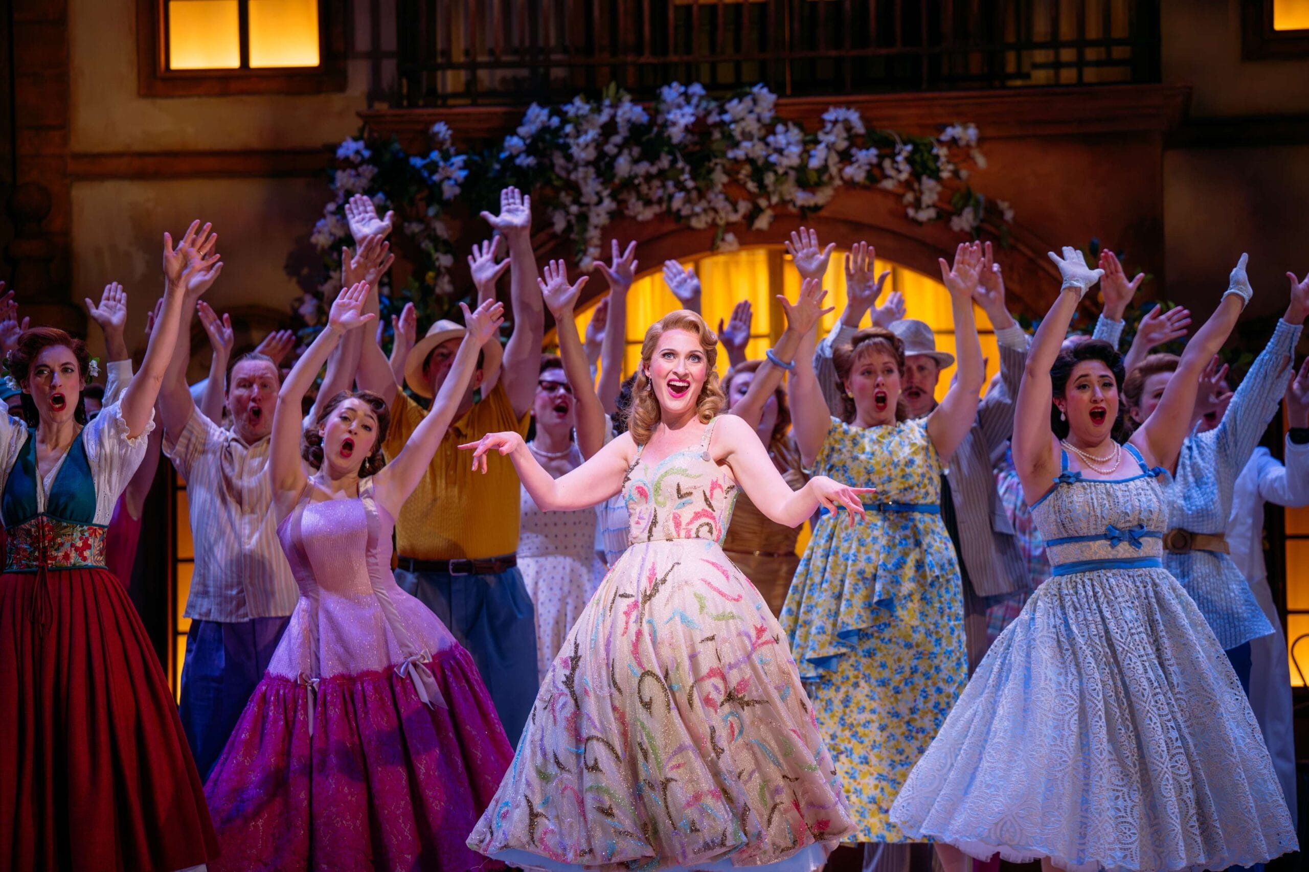 A group of performers in colorful vintage costumes sing and gesture with raised arms on a brightly lit stage decorated with flowers.