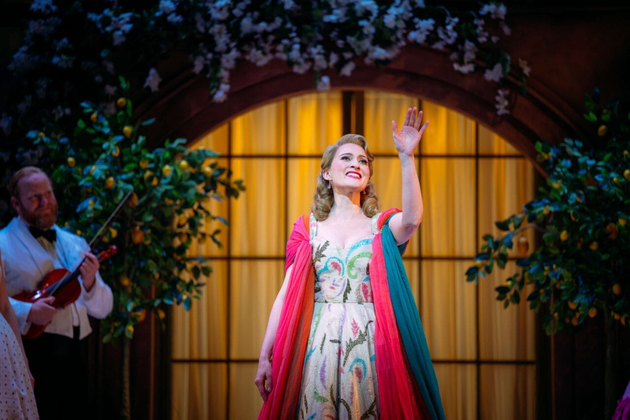 A woman in a colorful dress stands on stage, smiling and waving, with a violinist in the background and decorative plants and lights around her.