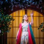 A woman in a colorful dress stands on stage, smiling and waving, with a violinist in the background and decorative plants and lights around her.