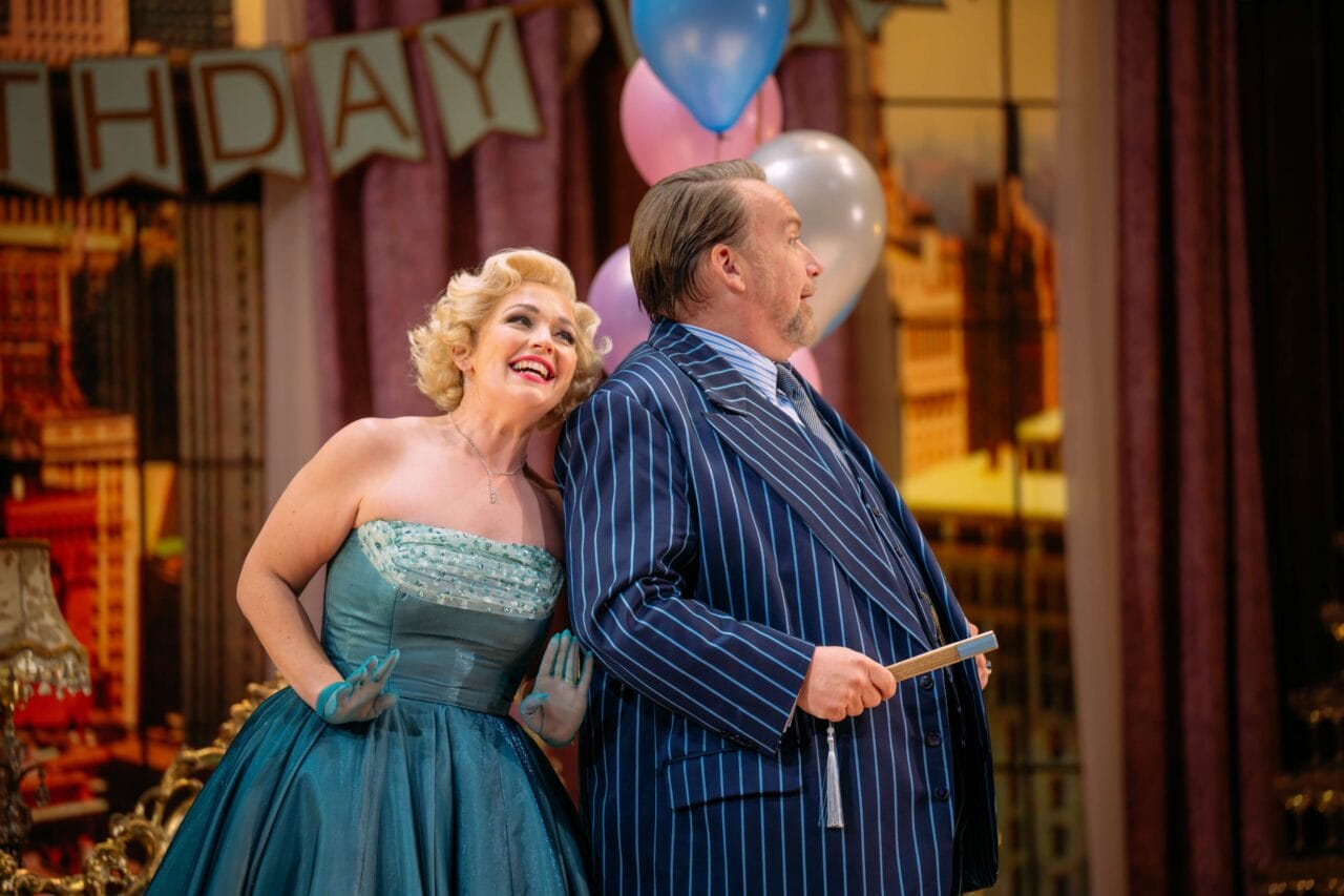 A woman in a teal strapless dress leans cheerfully against a man in a blue pinstripe suit; both stand in front of birthday decorations and balloons.