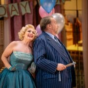 A woman in a teal strapless dress leans cheerfully against a man in a blue pinstripe suit; both stand in front of birthday decorations and balloons.