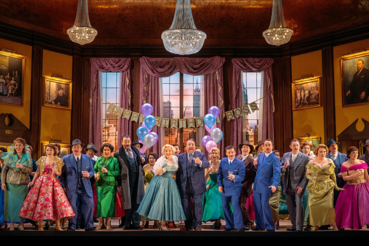 A group of people in colorful, vintage-style costumes stand in a decorated room with chandeliers, balloons, and a “Happy Birthday Boss” banner, appearing to celebrate an event.