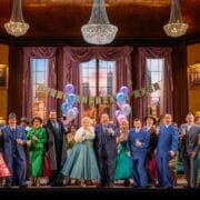 A group of people in colorful, vintage-style costumes stand in a decorated room with chandeliers, balloons, and a “Happy Birthday Boss” banner, appearing to celebrate an event.