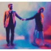 A man in a suit and a woman in a long skirt and coat stand holding hands; the image is blurred with a colorful, abstract background in blue, pink, and orange tones.