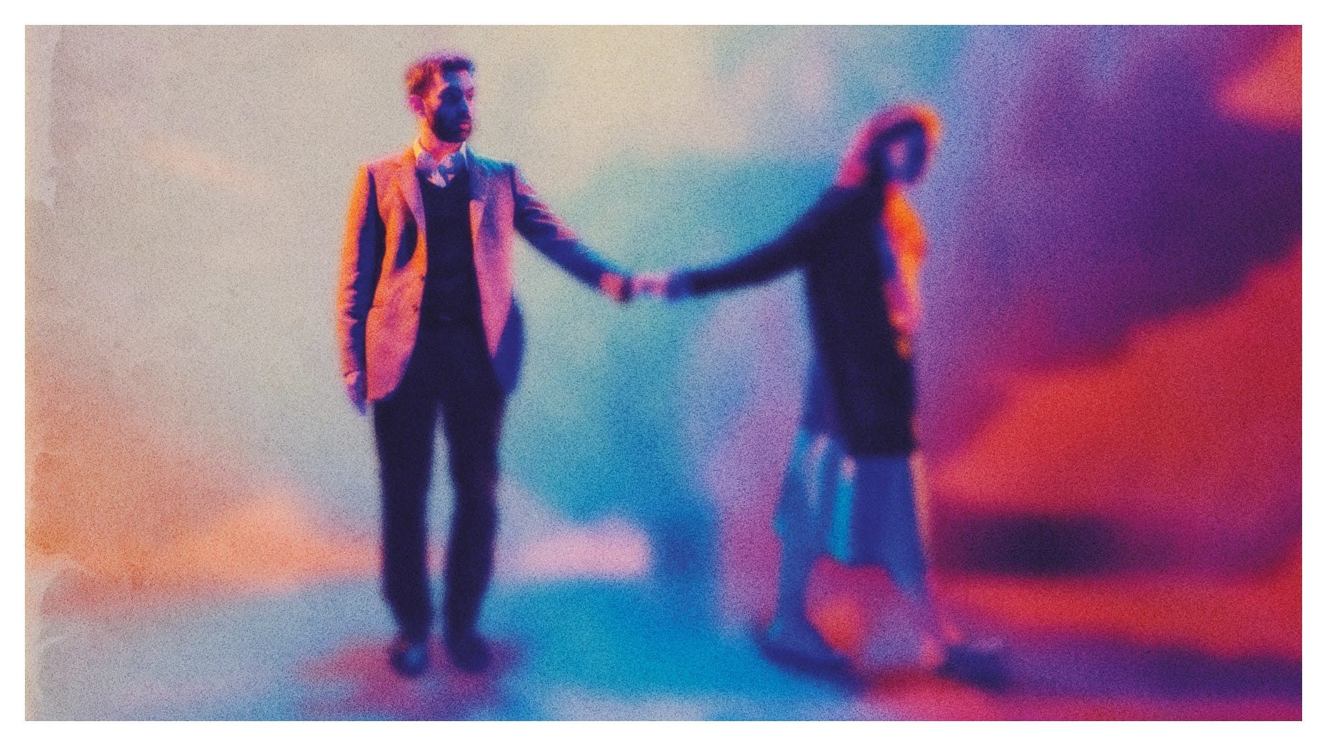 A man in a suit and a woman in a long skirt and coat stand holding hands; the image is blurred with a colorful, abstract background in blue, pink, and orange tones.