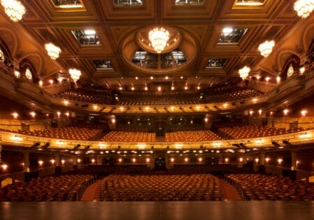 Interior of an empty, ornate theater viewed from the stage, featuring multiple levels of seating, decorative lights, and a central chandelier.
