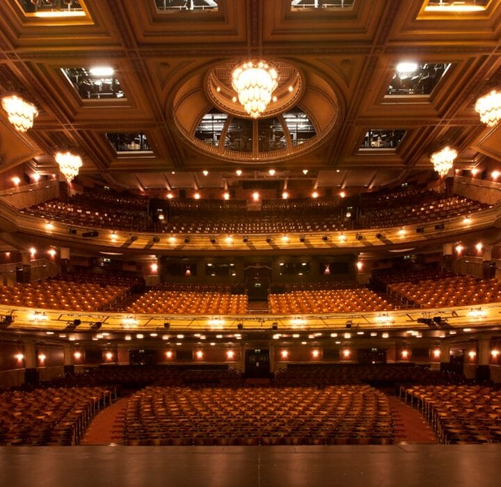 Interior of an empty, ornate theater viewed from the stage, featuring multiple levels of seating, decorative lights, and a central chandelier.