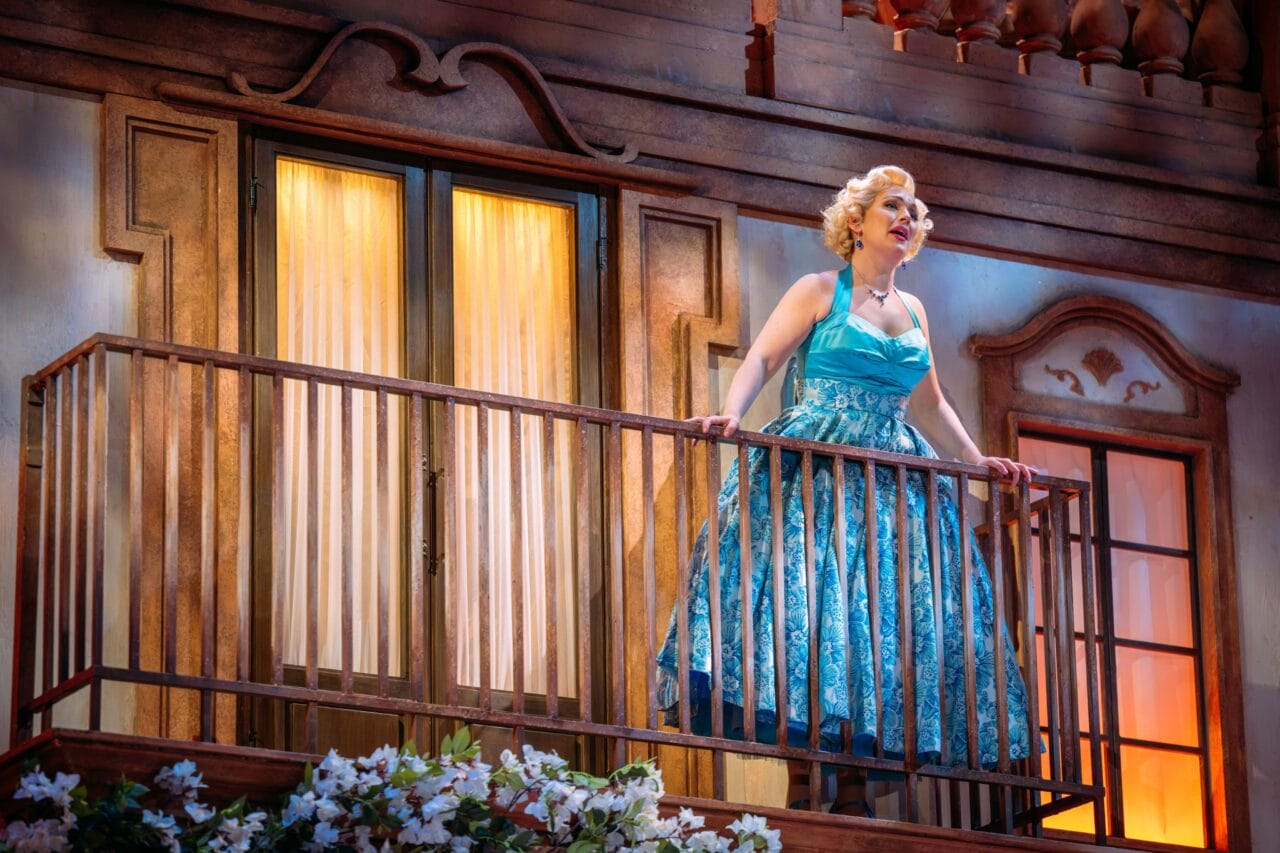 A woman in a blue patterned dress stands on a balcony in front of a building, appearing to sing or speak, with flowers visible below her.