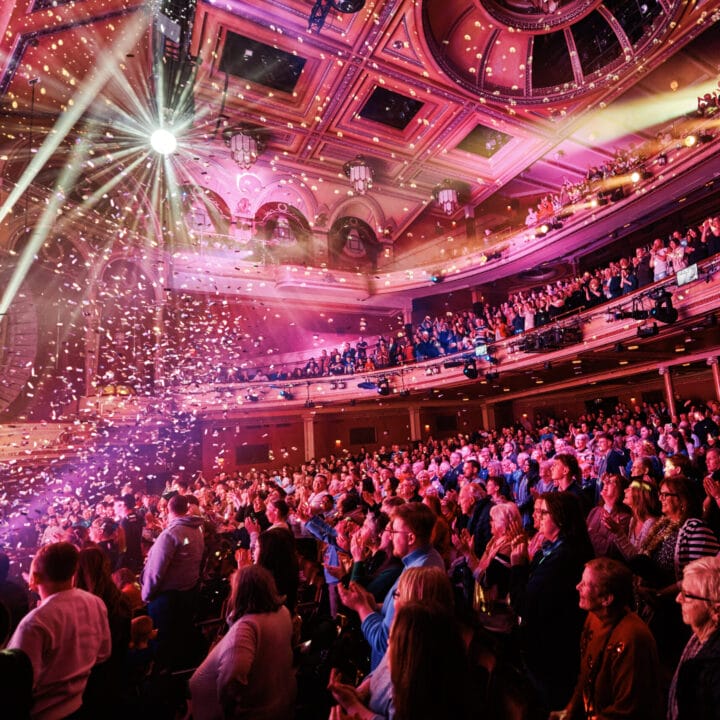 A large audience sits and stands in a grand theater, watching a brightly lit stage with confetti falling and colorful lights illuminating the ornate interior.