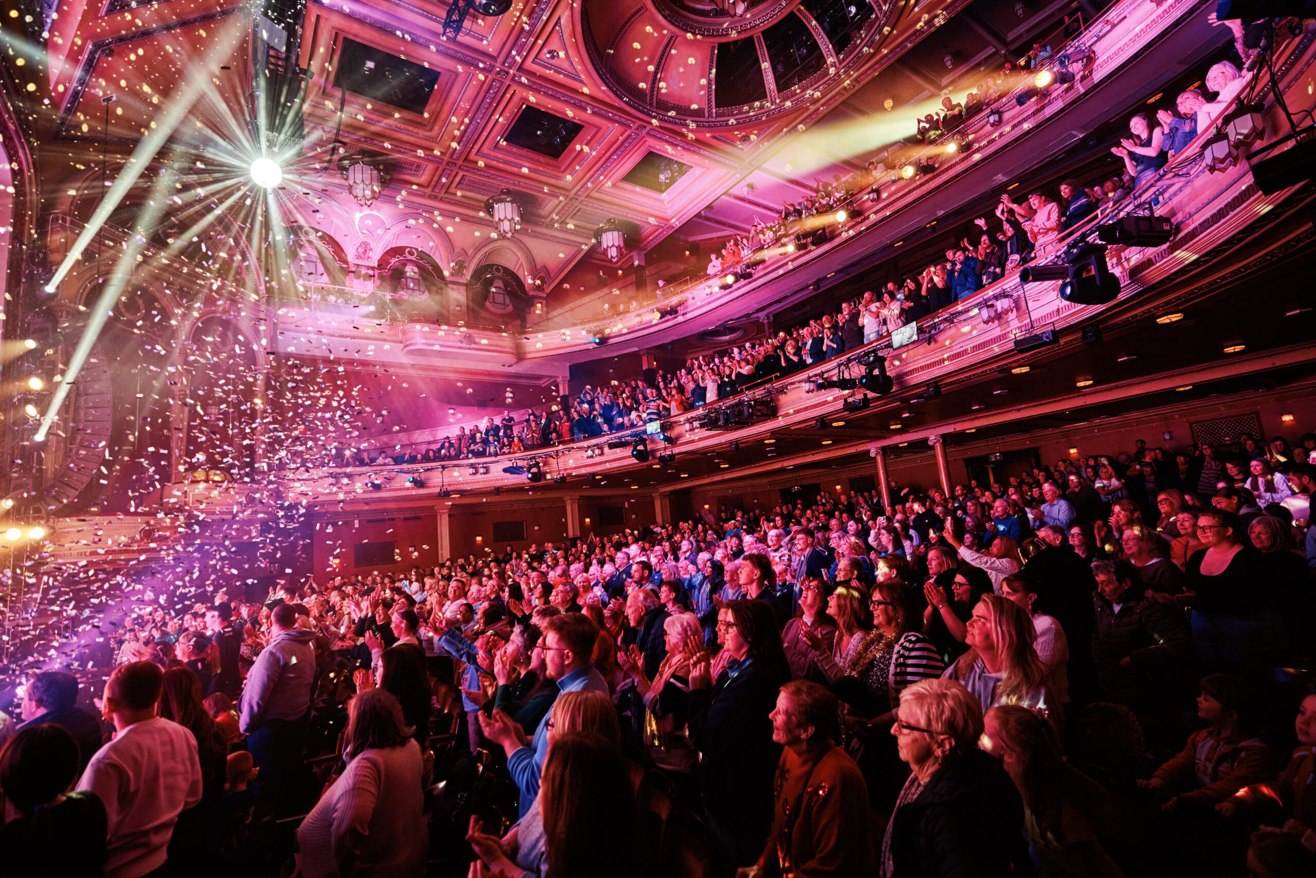 A large audience sits and stands in a grand theater, watching a brightly lit stage with confetti falling and colorful lights illuminating the ornate interior.