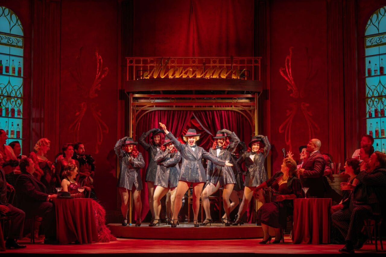 A group of performers in glittering costumes and top hats dance on stage under red lighting, surrounded by an audience seated at tables.