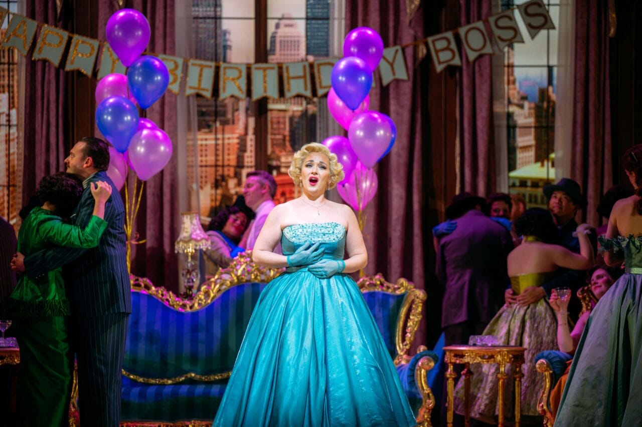 A woman in a turquoise gown sings at a lively indoor party decorated with purple balloons and a "Happy Birthday Boss" banner.
