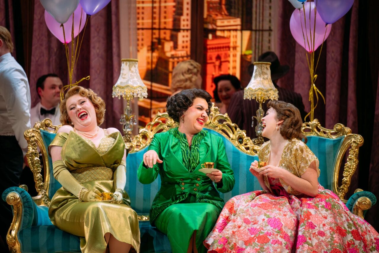 Three women in vintage dresses sit laughing on an ornate blue sofa at a lively party, holding drinks, with balloons and other guests in the background.