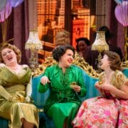 Three women in vintage dresses sit laughing on an ornate blue sofa at a lively party, holding drinks, with balloons and other guests in the background.