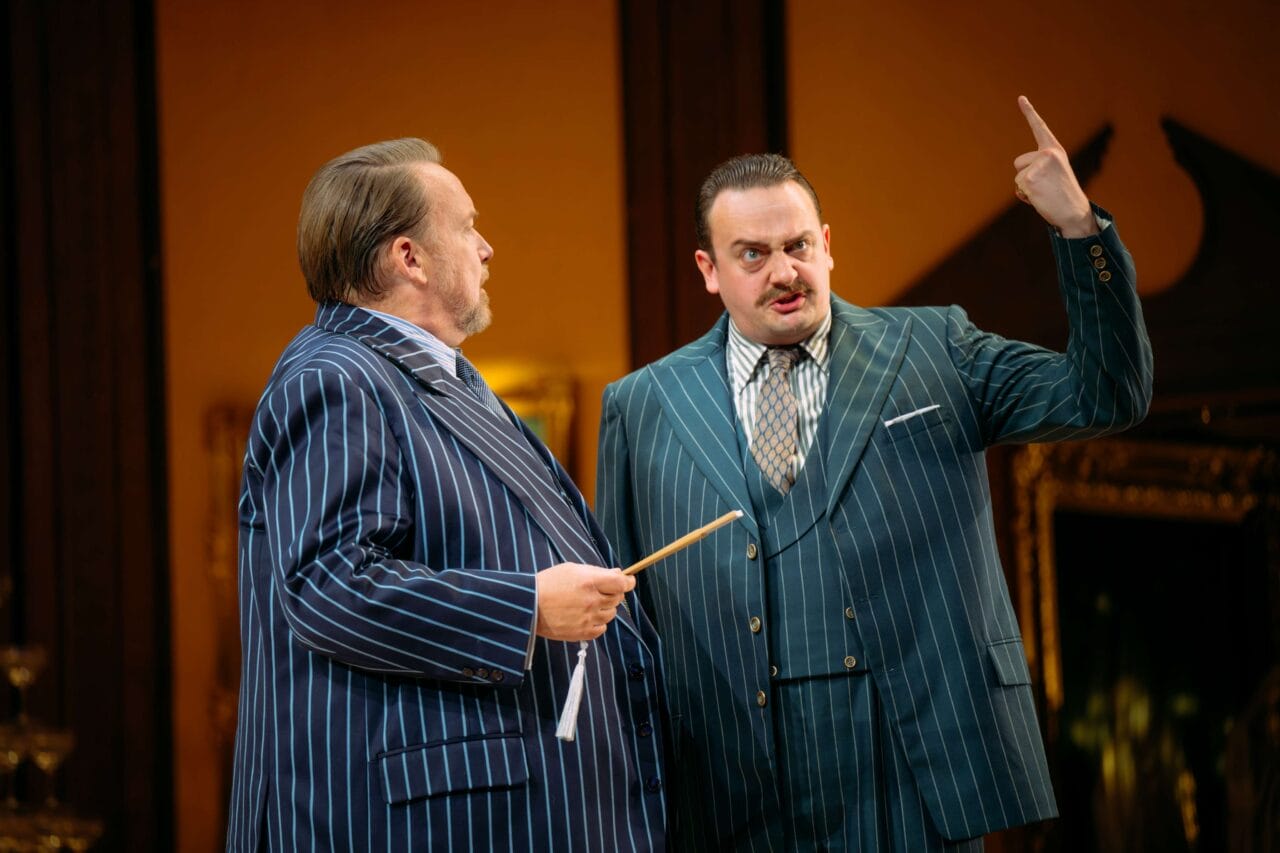 Two men in pinstripe suits stand indoors; one gestures with a raised finger while the other holds a stick, both appearing engaged in conversation.