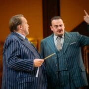 Two men in pinstripe suits stand indoors; one gestures with a raised finger while the other holds a stick, both appearing engaged in conversation.