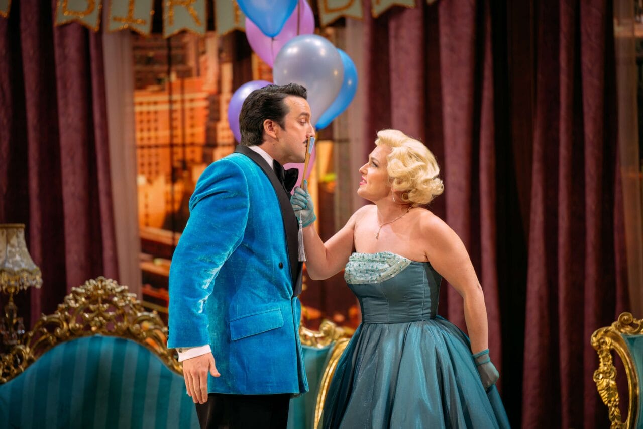 A man in a blue velvet jacket and a woman in a teal strapless dress stand close together in a lavishly decorated room, holding balloons and making eye contact.