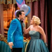 A man in a blue velvet jacket and a woman in a teal strapless dress stand close together in a lavishly decorated room, holding balloons and making eye contact.
