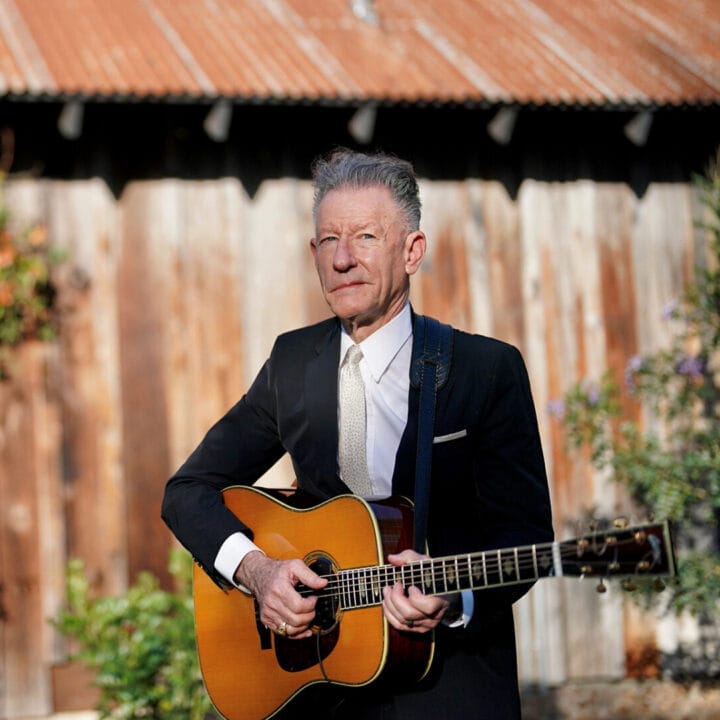 A man in a suit stands outdoors in front of a rustic wooden building, playing an acoustic guitar.