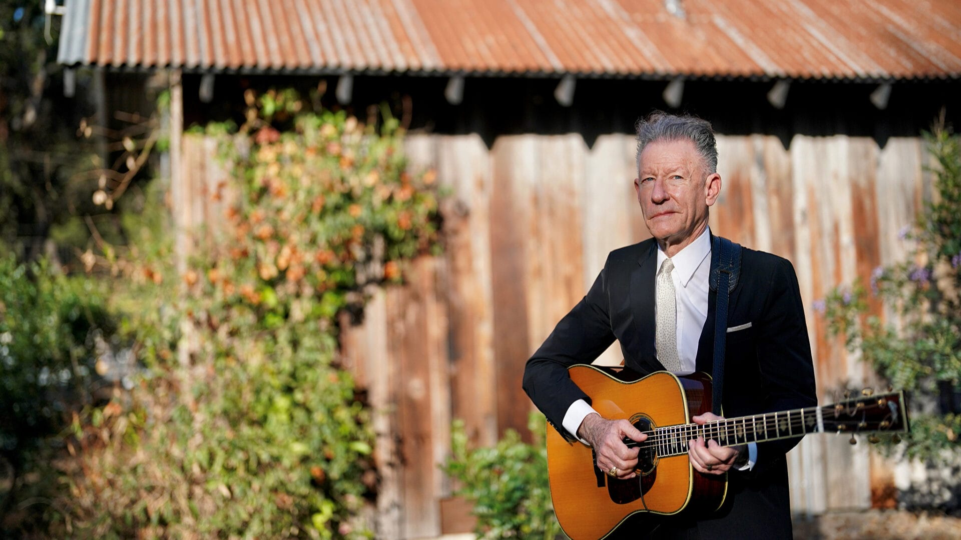 A man in a suit stands outdoors in front of a rustic wooden building, playing an acoustic guitar.