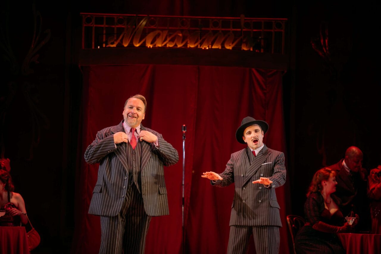 Two men in pinstripe suits perform on stage with a red curtain and neon sign in the background; one adjusts his jacket while the other gestures, with seated people visible at the sides.