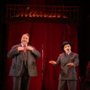 Two men in pinstripe suits perform on stage with a red curtain and neon sign in the background; one adjusts his jacket while the other gestures, with seated people visible at the sides.