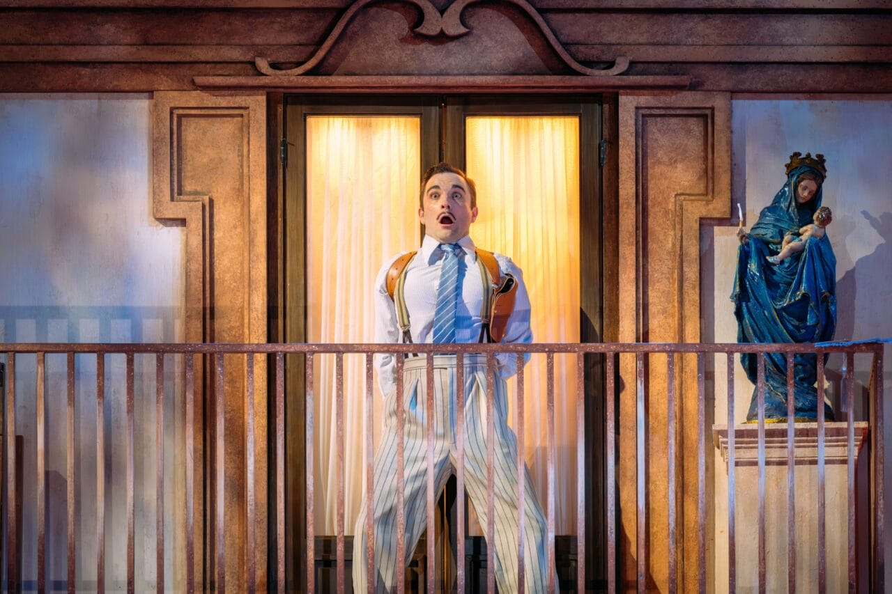 A man in striped pajamas stands surprised on a balcony in front of glass doors, with a statue of the Virgin Mary holding baby Jesus on a pedestal nearby.