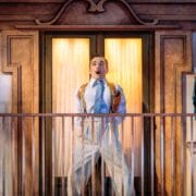 A man in striped pajamas stands surprised on a balcony in front of glass doors, with a statue of the Virgin Mary holding baby Jesus on a pedestal nearby.