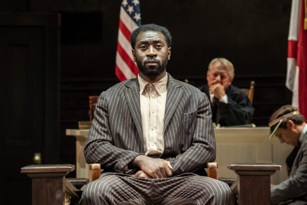 A man in a pinstripe suit sits in a courtroom witness stand, evoking scenes from Harper Lee’s classic novel To Kill a Mockingbird. A judge and two others are visible, with an American flag standing behind them.