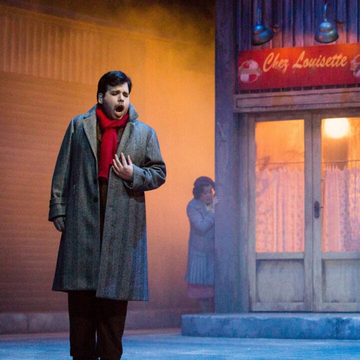 Two men stand onstage in front of a shop facade, dressed in winter clothing, with another person visible in the background.