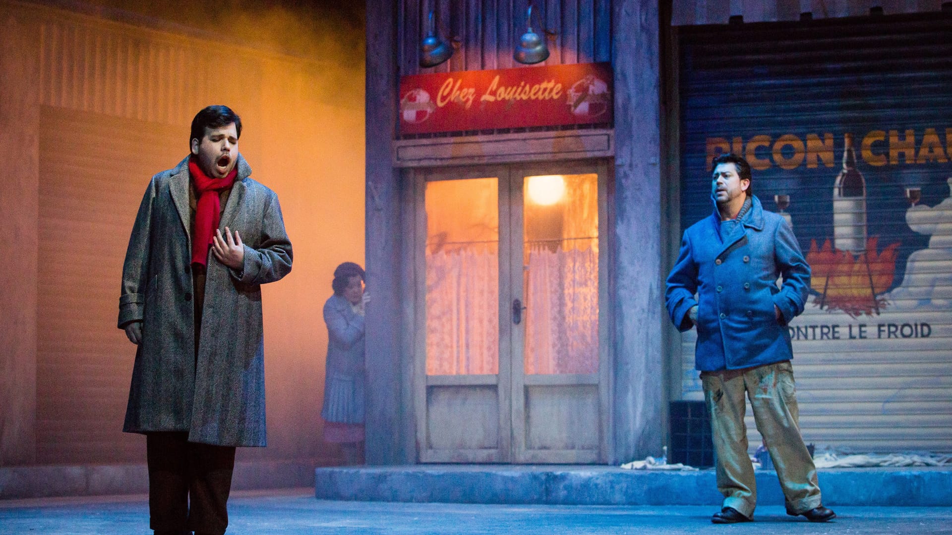 Two men stand onstage in front of a shop facade, dressed in winter clothing, with another person visible in the background.