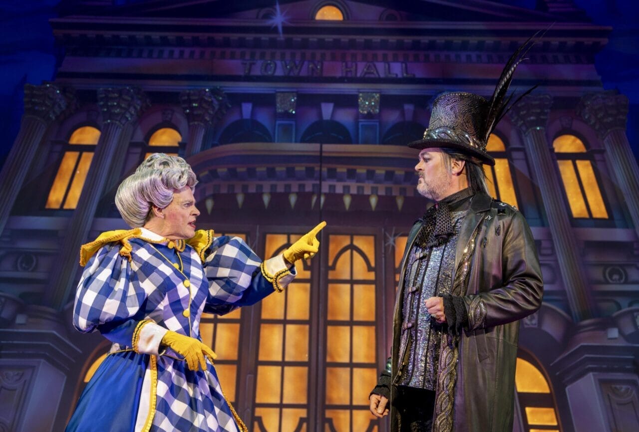 Two actors in theatrical costumes perform on stage; one in a blue plaid dress and gloves points at the other, who wears a black coat and tall hat, as a whimsical Jack and the Beanstalk town hall backdrop sets the scene behind them.