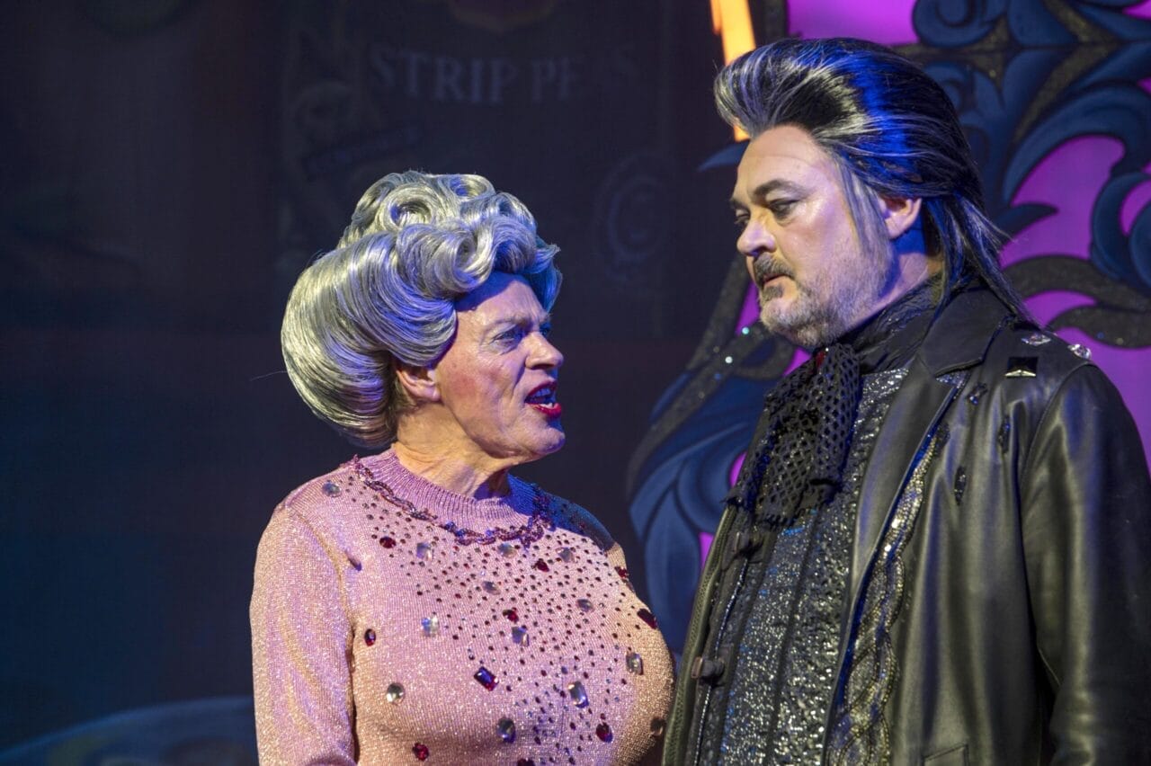 Two actors in elaborate costumes and wigs perform on stage in a lively Jack and the Beanstalk scene; one wears a jeweled pink sweater, while the other dons a dark coat with silver-streaked hair.