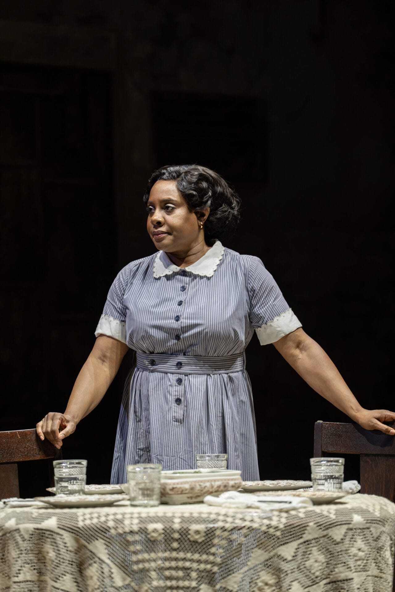 A woman in a striped dress with a white collar stands behind a table set with glasses and a lace tablecloth, evoking the timeless elegance of a classic novel like Harper Lee’s To Kill a Mockingbird, as she rests her hands on two chairs.