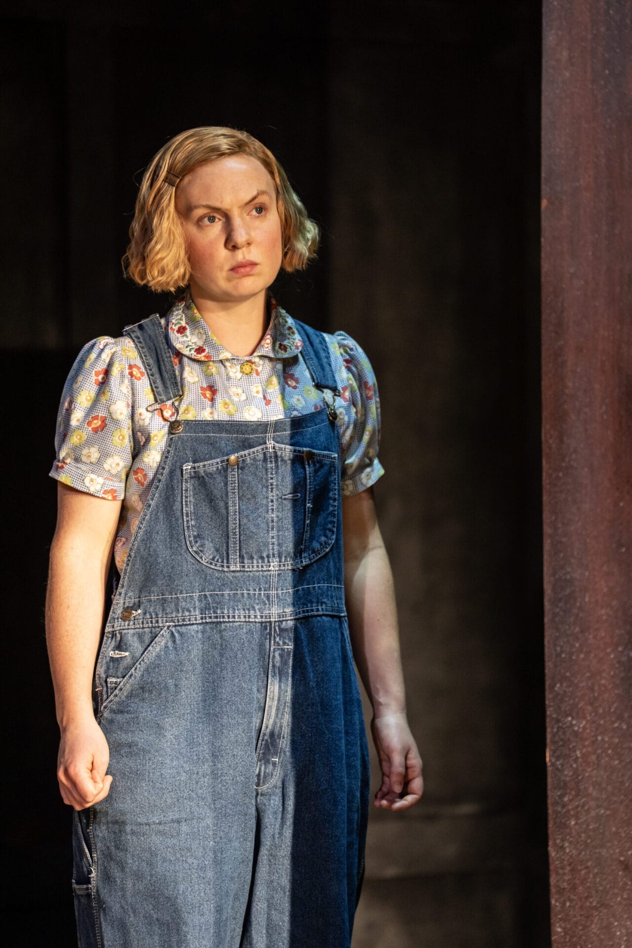 A woman with short blonde hair, wearing a floral shirt and blue denim overalls, stands indoors looking slightly to the side with a neutral expression—evoking the quiet strength of a character from a classic novel like To Kill a Mockingbird.