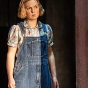 A woman with short blonde hair, wearing a floral shirt and blue denim overalls, stands indoors looking slightly to the side with a neutral expression—evoking the quiet strength of a character from a classic novel like To Kill a Mockingbird.