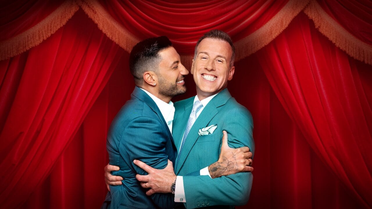 Two men in teal suits smile and embrace in front of red stage curtains.
