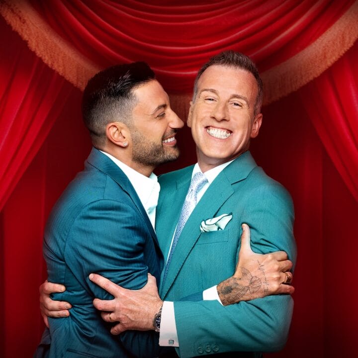 Two men in teal suits smile and embrace in front of red stage curtains.
