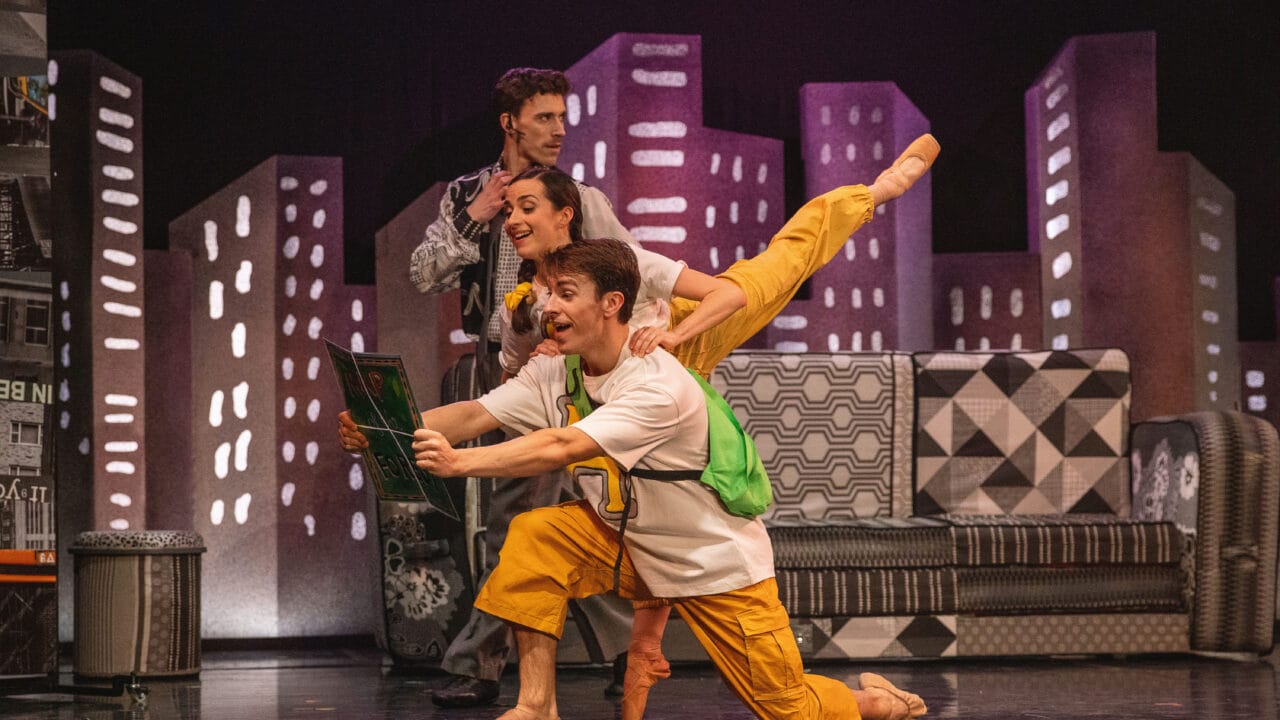 Three performers in colourful costumes pose energetically on stage with a cityscape backdrop, one holding a framed picture, while another balances on one leg behind him.