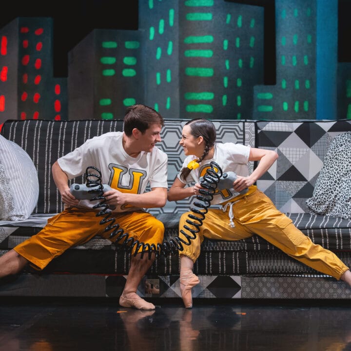 Two dancers in matching yellow pants and white shirts sit on a patterned couch, holding coiled cords and facing each other in a dynamic, mirrored pose.