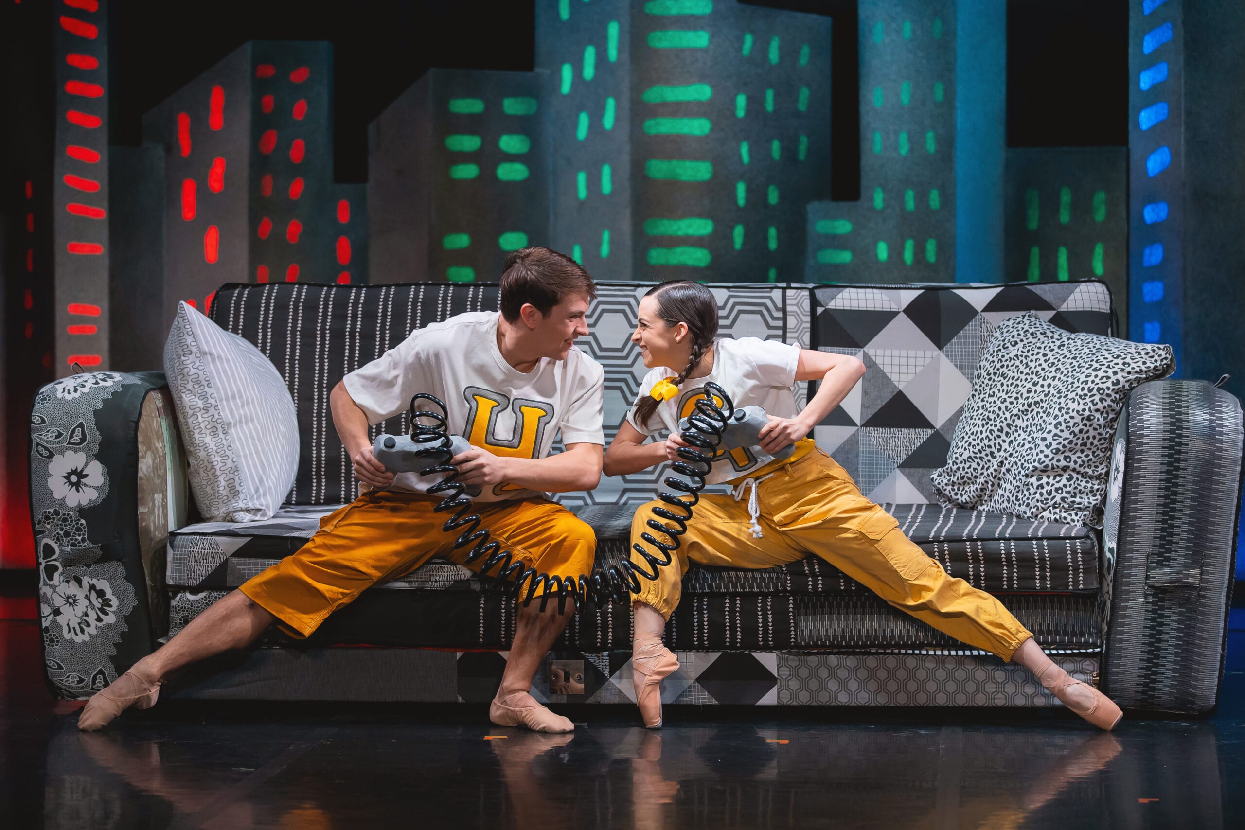 Two dancers in matching yellow pants and white shirts sit on a patterned couch, holding coiled cords and facing each other in a dynamic, mirrored pose.