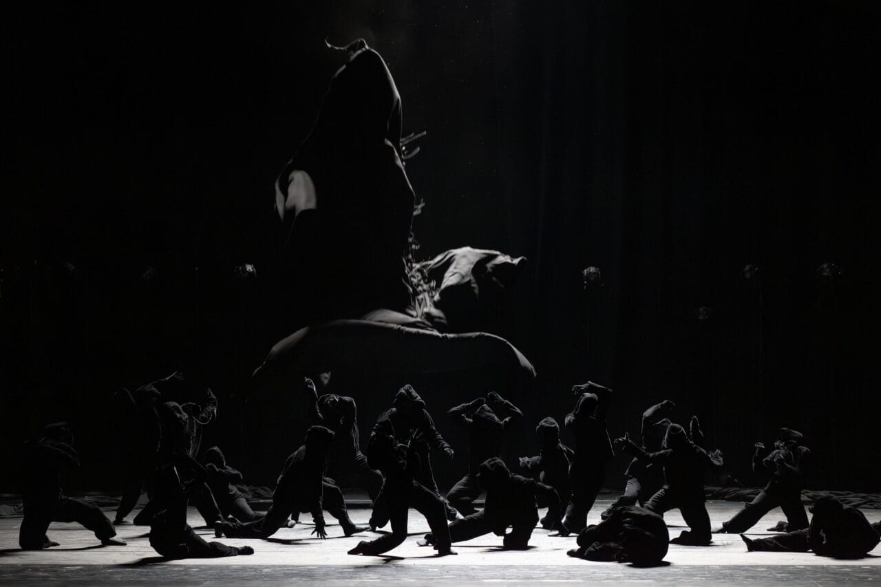 A group of dancers in dark costumes perform on a dimly lit stage, with one dancer leaping high above the others in the background.