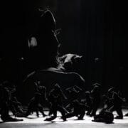 A group of dancers in dark costumes perform on a dimly lit stage, with one dancer leaping high above the others in the background.