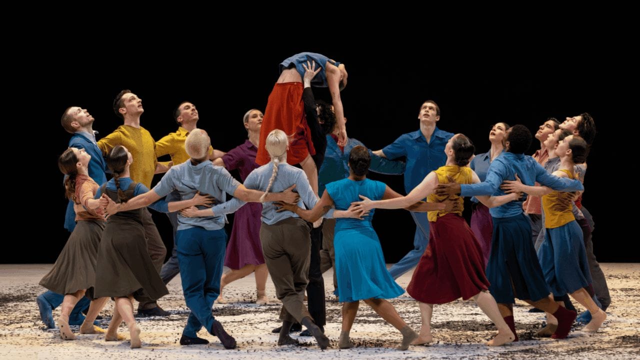 A group of dancers in colorful costumes form a circle on stage, lifting one dancer above their heads during a performance.