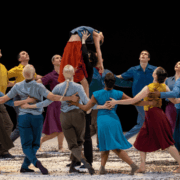 A group of dancers in colorful costumes form a circle on stage, lifting one dancer above their heads during a performance.