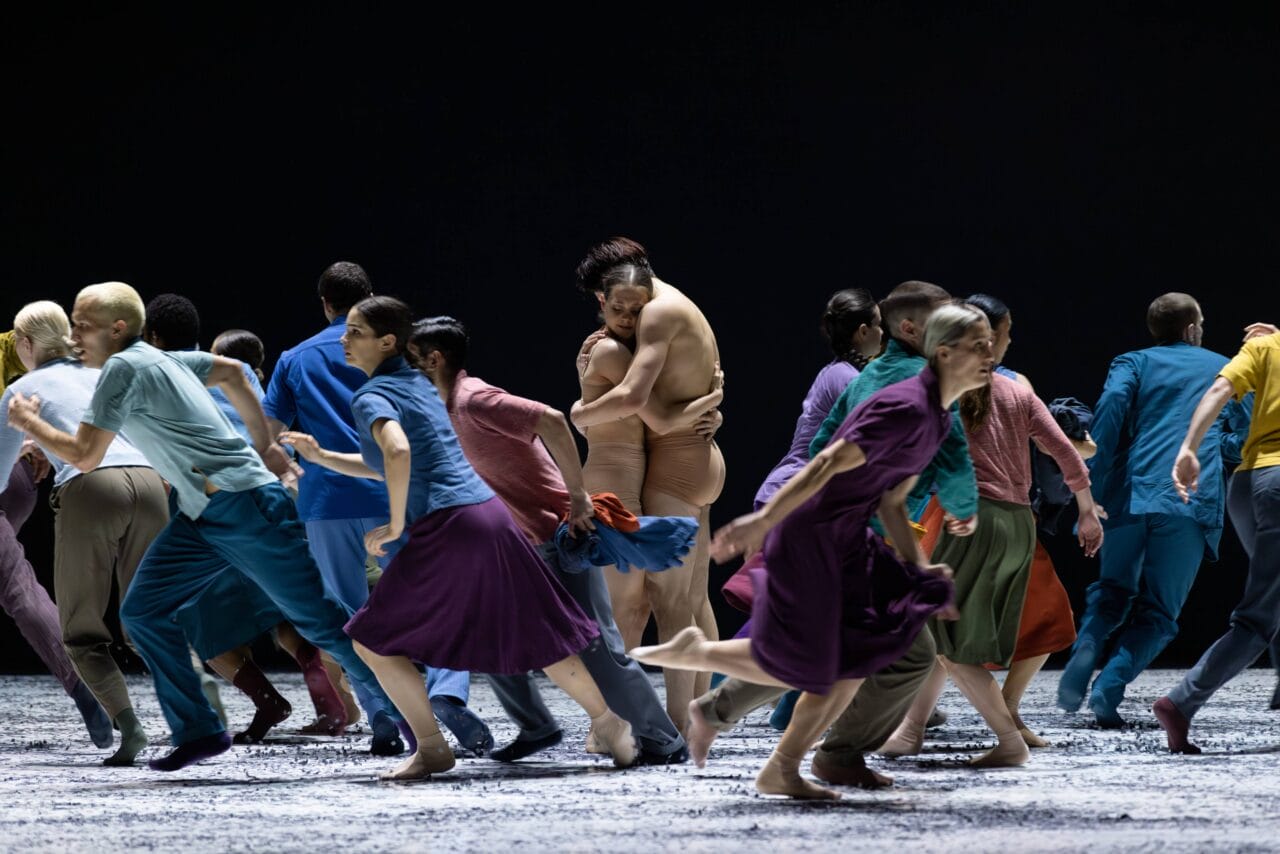 A group of dancers in colorful clothing move energetically on stage while two dancers, one in nude attire, embrace at the center.
